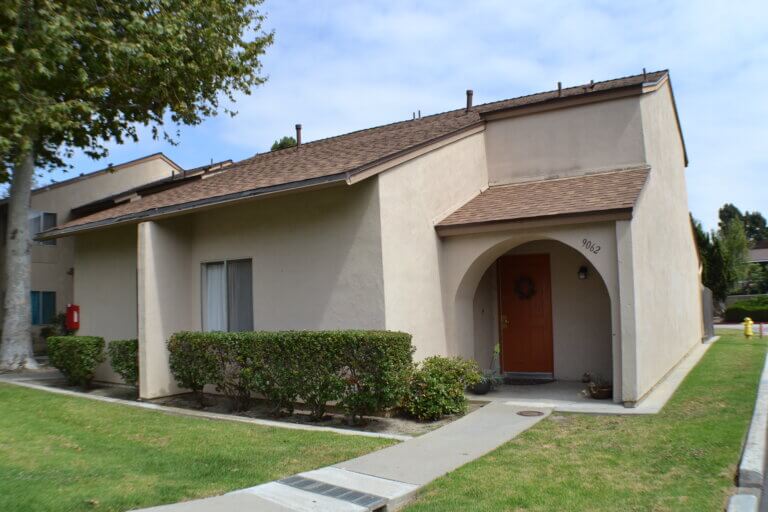 Corner unit with tan stucco, arched porch, front door, trimmed hedges, and sidewalk.