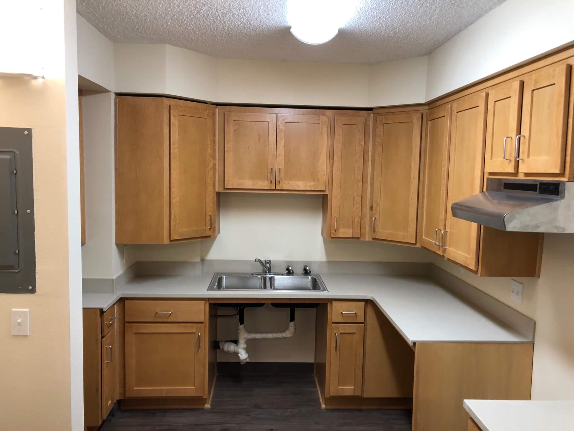 Compact U-shaped kitchen with light wood shaker cabinets, counters, double sink, and range hood.