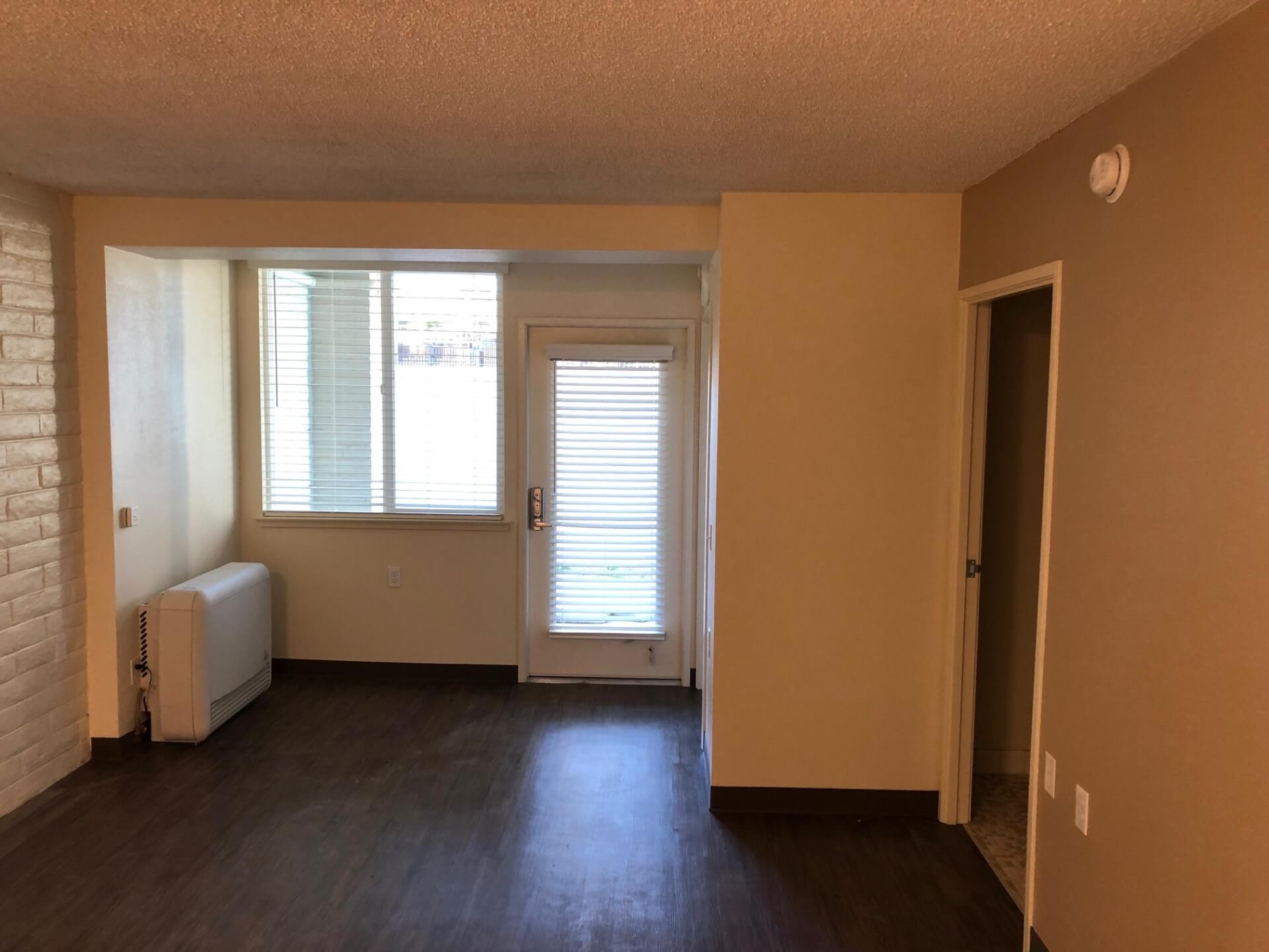 Living room with blinds, glass door to outside, wood flooring, and wall heater.