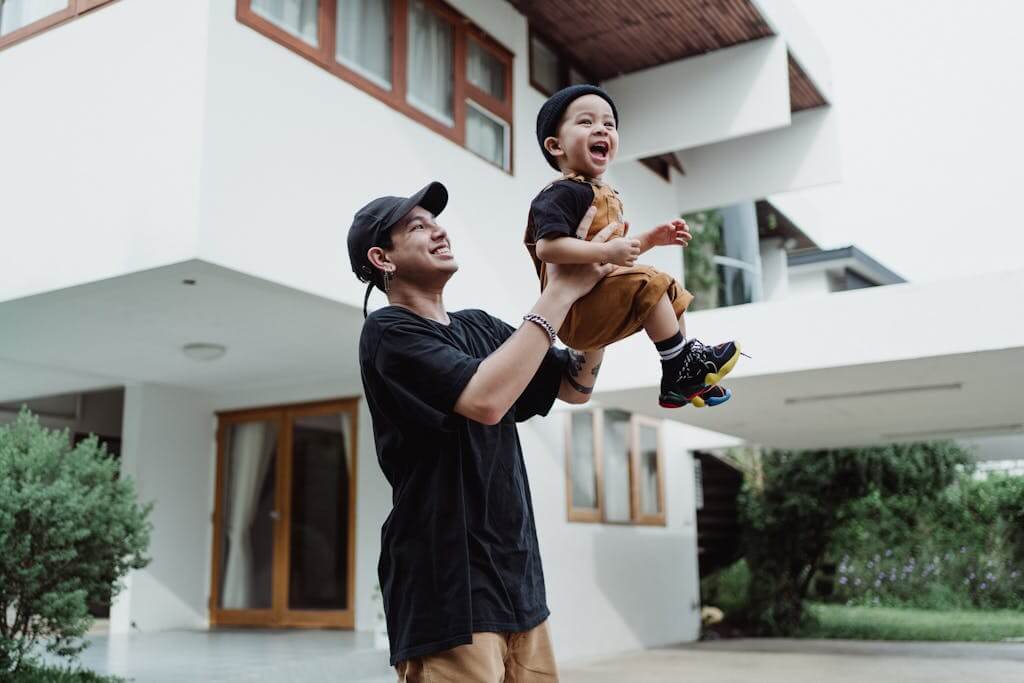 Father and son bonding outside modern house during daytime, enjoying happy playful lifting.